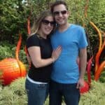 Couple standing close together outdoors, woman in black shirt and jeans and man in blue shirt smiling, with orange glass sculpture art behind them.