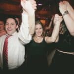 Friends at a party raise their hands together in celebration, smiling at the camera, close to the foreground.