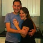 A smiling couple hugging in a modern kitchen, the man in a light blue shirt and the woman in black.