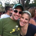A smiling couple posing for a close-up photo at an outdoor event, with people and trees in the background.