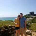 Couple standing together outdoors by the coast, with ruins and blue ocean in the background, smiling at the camera.