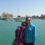 Couple standing by a turquoise harbor, smiling, with boats and a city skyline in the background.