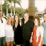 Six adults standing arm-in-arm and smiling outdoors near a pool, with palm trees in the background.