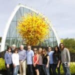 Group of twelve people posing in front of a large yellow Chihuly glass sculpture beside a glass-walled conservatory at Chihuly Garden and Glass, with greenery around.