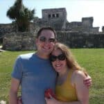 Smiling couple wearing sunglasses stands close together in front of an ancient stone ruin on a sunny day.