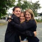 Three people wearing dark jackets hug and smile outdoors by a waterfront marina, with a camera visible in one person's hand.