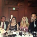 Group of people in formal attire dining at a long table with wine glasses, napkins, and flowers; a lit green EXIT sign is visible on the wall behind them.