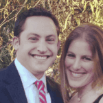 Smiling couple in formal attire posing together outdoors with green foliage in the background.
