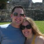 Smiling couple wearing sunglasses hugs in front of stone ruins on a sunny day.